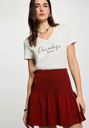 Donna che indossa una maglietta bianca con scollo a V con la scritta «Paradise Island» e una collana a forma di cuore, abbinata a una gonna rossa a vita alta con orlo arricciato.