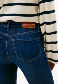 Isik, kes kannab tumedates sinistes teksades nahast Tommy Hilfigeri sildiga vöökohal ning valget triibulist pika varrukaga särk mustade horisontaalsete triipudega.