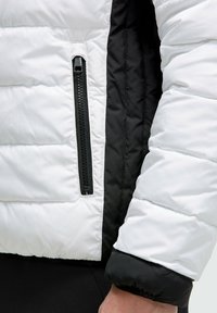 Chaqueta tipo pluma con un exterior blanco y acentos negros. Cuenta con un bolsillo con cremallera y un diseño de paneles distintivo. Textura suave, aislante.