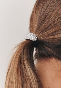 Schwarzes Haargummi mit silberner, strassverzierter Dekoration, das einen mittellangen braunen Pferdeschwanz sicher hält.