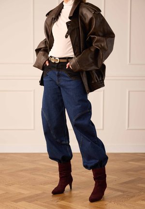 Person wearing dark brown oversized leather jacket, white turtleneck, loose blue jeans, black belt, and burgundy heeled boots.