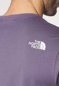 Fialové bavlněné tričko s kulatým výstřihem a krátkým rukávem, na horní části zad s bílým logem. Hladká textura.