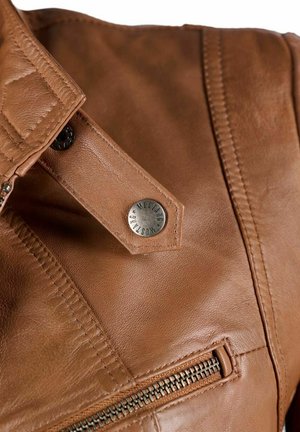 Nahaufnahme einer braunen Lederjacke mit Metallsnap-Knopf mit dem Text "Mustang" und einer Reißverschlusstasche.