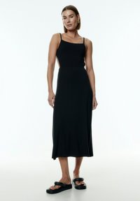 Schwarzes Midikleid mit Spaghettiträgern, taillierter Bustier und seitlichen Ausschnitten. Glatte Textur und fließende Silhouette. Getragen mit klobigen schwarzen Sandalen.