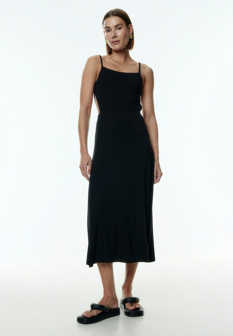 Schwarzes Midikleid mit Spaghettiträgern, taillierter Bustier und seitlichen Ausschnitten. Glatte Textur und fließende Silhouette. Getragen mit klobigen schwarzen Sandalen.