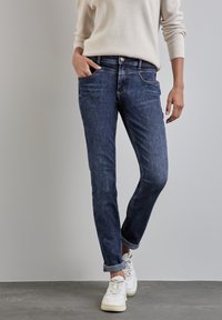 Jean en denim bleu foncé avec une coupe slim, un design à cinq poches et des ourlets retournés, associé à des baskets blanches et un pull beige clair.