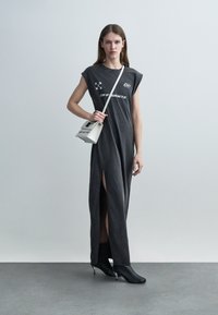 Młoda kobieta w czarnej, bezrękawnikowej sukience maxi z nadrukami logo, czarnych botkach na obcasie i małej białej torebce na ramię na szarym tle.