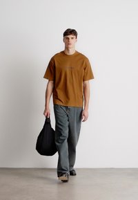 Καφέ βαμβακερό μπλουζάκι με στρογγυλή λαιμόκοψη και κοντά μανίκια, σε συνδυασμό με γκρι τζιν παντελόνι. Μια μαύρη τσάντα ώμου είναι κρεμασμένη στον ώμο.