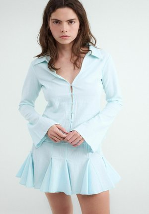 Mladá žena s dlhými hnedými vlasmi, ktorá má na sebe svetlo modrú gombíkovú blúzku s rozšírenými rukávmi a zodpovedajúcu plisovanú sukňu, stojaca pred jednoduchým pozadím.