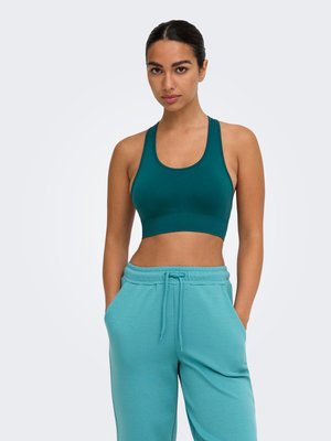 Femme aux cheveux foncés attachés, portant un brassière de sport turquoise et un pantalon de survêtement bleu clair, mains dans les poches, sur un fond uni.
