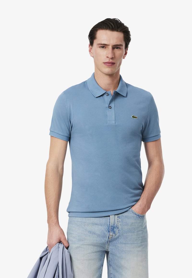 Polo de color azul claro hecho de algodón, con un cuello clásico, un cierre de dos botones y un pequeño logo verde en el lado izquierdo del pecho. Llevado con jeans claros.