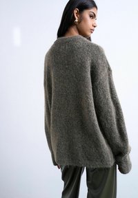 Jeune femme portant un pull gris pelucheux surdimensionné et un pantalon foncé, vue de profil sur un fond uni clair.