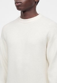 Gros plan d'une personne portant un pull blanc uni à col rond, montrant le haut de la poitrine, le cou et la partie inférieure du visage.