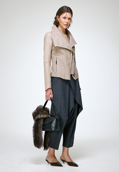 Femme portant une veste beige avec des manches en cuir, un pantalon ample gris foncé, des talons noirs pointus, tenant un sac à main noir avec un accessoire en fourrure.