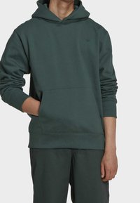 Man som bär en mörkgrön huvtröja med framficka och matchande byxor, handen i fickan, mot en enkel ljus bakgrund.