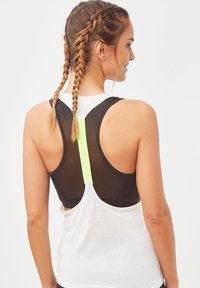 Mujer con dos trenzas lleva una blusa sin mangas blanca sobre un sujetador deportivo negro con una franja vertical de color amarillo neón en la parte posterior.