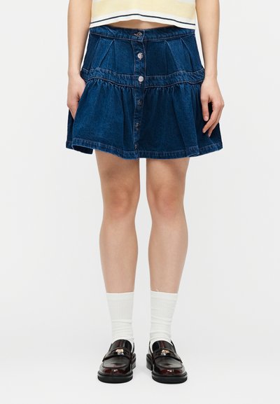 Άτομο που φοράει μπλε τζιν φούστα με κουμπιά μπροστά, λευκές κάλτσες και σκούρα καφέ μοκασίνια, στέκεται μπροστά σε απλό λευκό φόντο.