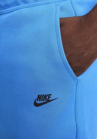 Mörkhyad hand i fickan på ljusblå Nike-träningsbyxor, med svart Nike-logotyp på tyget.