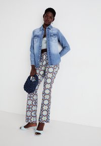 Camisa de denimb com bolsos pontiagudos, calças largas estampadas em azul, amarelo e flores castanhas, saltos azuis e uma mala de textura azul-marinho.