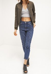 Oliwkowa bomberka z teksturowanej tkaniny, szary krótki top oraz niebieskie dżinsy. Czarne sandały na obcasie, swobodny krój, podwinięte mankiety.