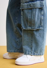 Pantaloni cargo in denim azzurro chiaro con grandi tasche laterali, abbinati a sneakers bianche con suola viola e dettagli perforati.