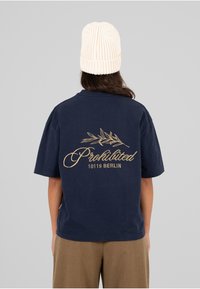 Camiseta de algodón azul marino con letras doradas "Prohibido 10119 BERLÍN" y un diseño de rama de olivo en la parte posterior, corte relajado.