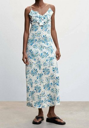 Femme portant une longue robe blanche sans manches avec un imprimé floral bleu et un col à volants, associée à des sandales compensées noires.