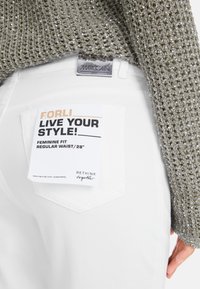 Weiße Jeans mit normaler Taille, versehen mit einem Aufnäher an der hinteren Tasche, der "Forli" und "Live Your Style!" in fettem Text zeigt. Glänzendes, glitzerndes grünes Stricktop.
