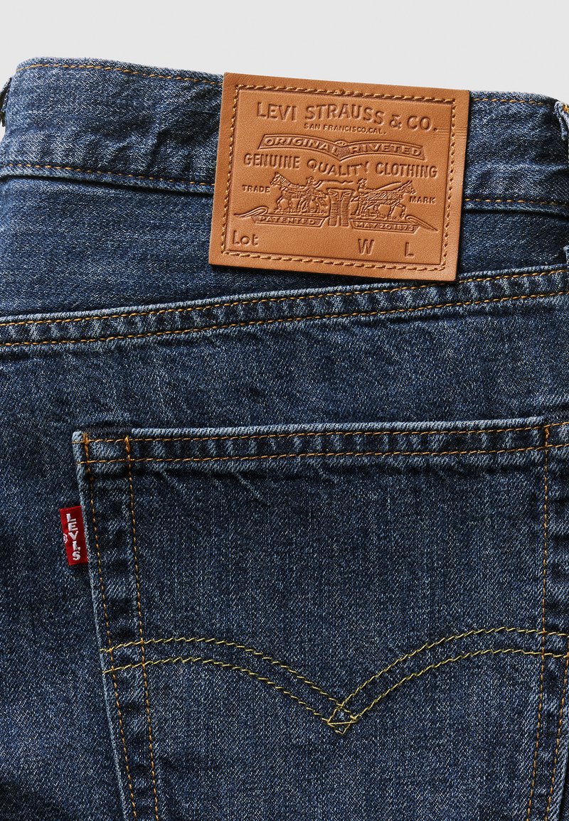 Jeans en denim bleu foncé avec une pièce en cuir tan portant un logo en relief au niveau de la taille. Coutures jaunes le long des poches et des coutures. Étiquette de logo rouge.