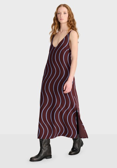 Femme aux longs cheveux roux portant une robe bordeaux sans manches avec des rayures ondulées bleues et des bottes noires, debout devant un fond uni.