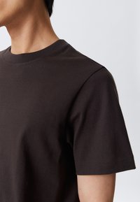 Personne portant un t-shirt à col rond à manches courtes de couleur marron foncé uni, vu de l'épaule au cou sur un fond clair.