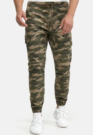 Mann iført grønne og brune kamuflasje cargo-bukser med elastiske mansjetter og hvite joggesko, stående nøytralt på hvit bakgrunn.