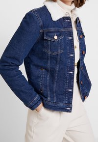 Person klädd i en blå jeansjacka med vit sherpa-krage och beige byxor, med händerna i fickorna mot en enhetlig bakgrund.