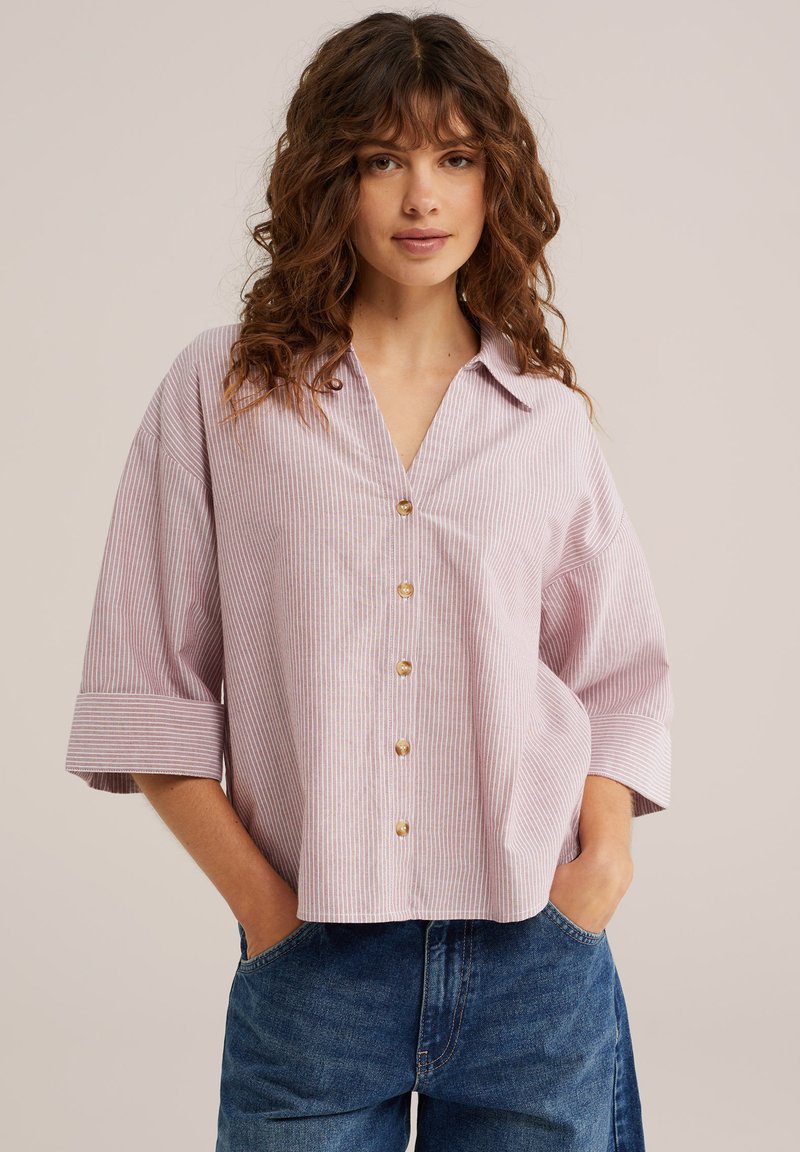 Woman with curly brown hair wearing a loose, light pink striped button-up shirt and blue jeans, hands in pockets, neutral background.