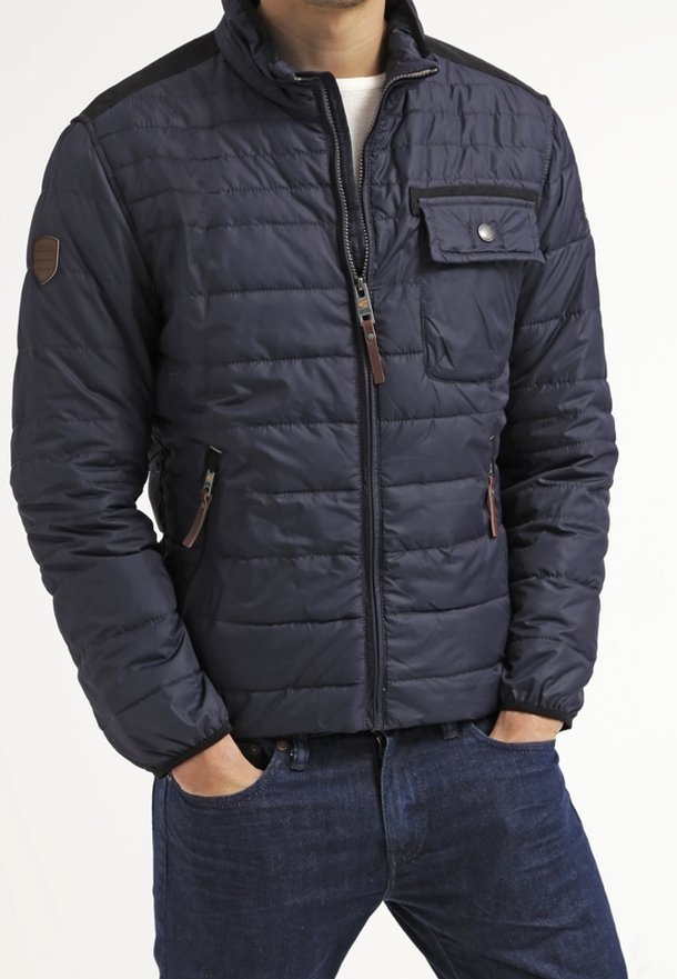 Uomo che indossa una giacca imbottita blu navale con chiusura frontale a zip, tasca sul petto con pattina e tasche laterali con zip, abbinata a jeans blu scuro.