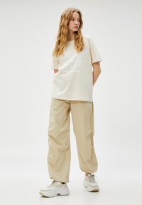 Beige bomulds t-shirt med et sommerfugle-grafik og tekst, parret med løstsiddende beige bukser og hvide sneakers med grå detaljer.