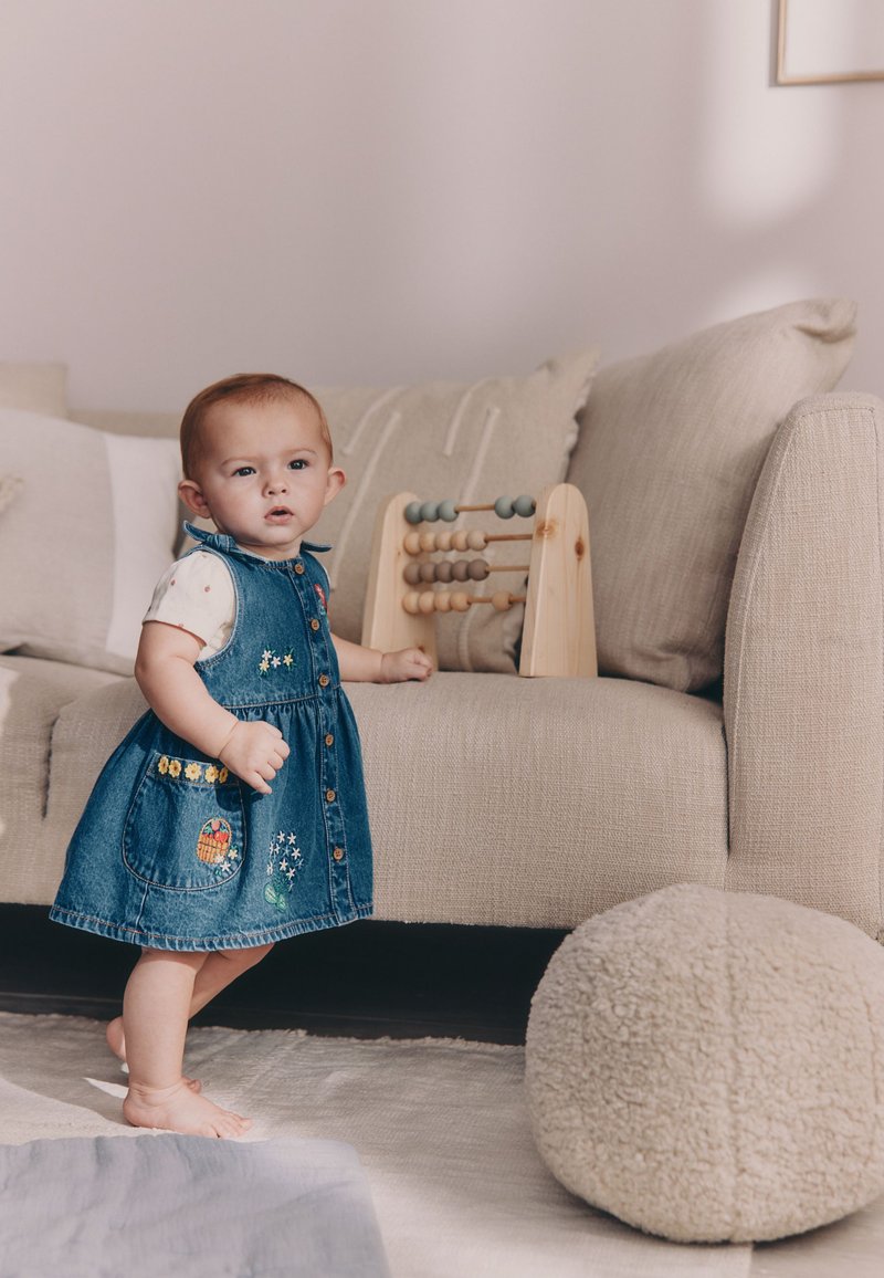 Toddler in denim dress standing barefoot near beige sofa with a wooden abacus toy and a round textured cushion on the floor.