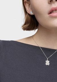Srebrny zestaw biżuterii z kolczykami w kształcie niedźwiedzia oraz pasującym wisiorkiem, oba o błyszczącym wykończeniu, noszone na ciemnej, teksturowanej bluzce.