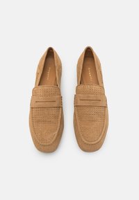 Mocassins en suède beige avec des détails perforés, une sangle surélevée à l'avant et une conception à bout arrondi. Semelle plate avec des accents minimalistes.