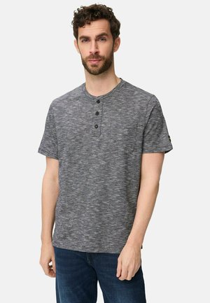 Mand med krøllet brunt hår og skæg iført en grå henley-t-shirt med korte ærmer og blå jeans, stående foran en ensfarvet baggrund.