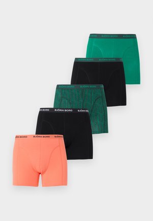 Μυώδης άνδρας όρθιος ξυπόλυτος μπροστά σε απλή λευκή επιφάνεια, φοράει κοραλλί μπόξερ Björn Borg, κοιτώντας μπροστά με χαλαρή στάση.