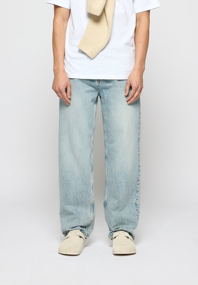 Personne portant un jean large bleu clair, un t-shirt blanc, un pull beige posé sur les épaules et des chaussures écru sans lacets, visible des épaules vers le bas.