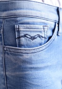 Jeans in denim bluastro chiaro con una tasca posteriore caratterizzata da cuciture in blu scuro a zigzag. La texture è liscia con una leggera scoloritura.