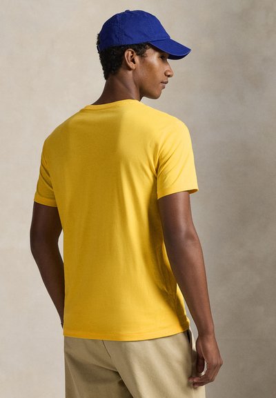 Κίτρινη βαμβακερή μπλούζα με κοντά μανίκια, στρογγυλή λαιμόκοψη, ομαλή υφή, σε συνδυασμό με μπλε καπέλο. Το μοντέλο είναι γυρισμένο με την πλάτη στην κάμερα.