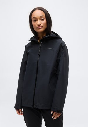 Γυναίκα με μαύρο μπουφάν με κουκούλα και φερμουάρ, μακρυμάνικο, στέκεται μπροστά σε λευκό απλό φόντο.