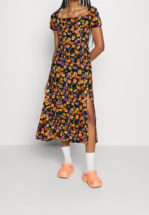Kvinde iført en sort midi-kjole med orange, lilla og gul blomsterprint, sideliste, hvide sokker og ferskenfarvede slip-on sko.