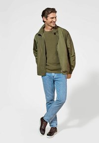 Lekka oliwkowa kurtka na ciemnozielonym swetrze wełnianym, zestawiona z niebieskimi dżinsami i brązowymi sznurowanymi trampkami.