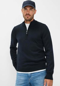 Maglione blu navy con zip e colletto a costine, realizzato in materiale lavorato a maglia. Indossato sopra una camicia bianca e abbinato a jeans azzurri chiari.