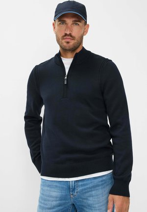 Marineblauer Zip-Kragen-Pullover aus Strickmaterial, mit einem gerippten Kragen und Saum. Getragen über einem weißen Hemd, kombiniert mit hellblauen Jeans.