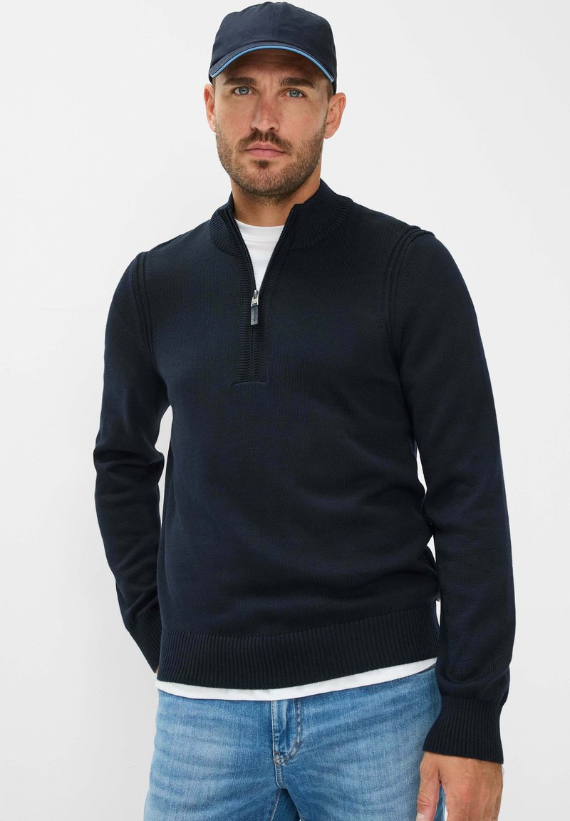 Maglione blu navy con zip e colletto a costine, realizzato in materiale lavorato a maglia. Indossato sopra una camicia bianca e abbinato a jeans azzurri chiari.